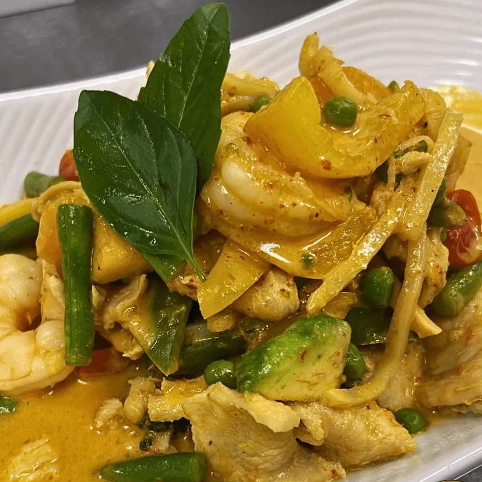 Close-up of a Thai curry dish with chicken, shrimp, vegetables, and basil on a white plate.