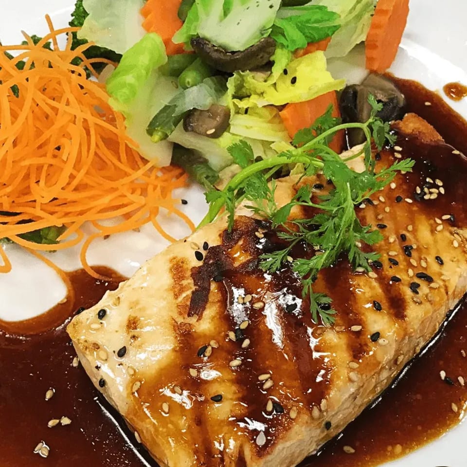 Grilled salmon with teriyaki sauce, sesame seeds, and a side of mixed vegetables and shredded carrots.