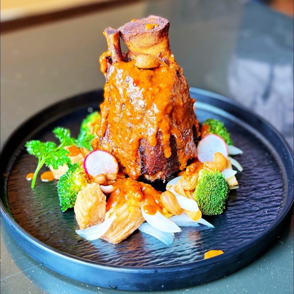 Braised pork hock with red curry sauce, broccoli, radishes, cashews, and onions on a black plate.